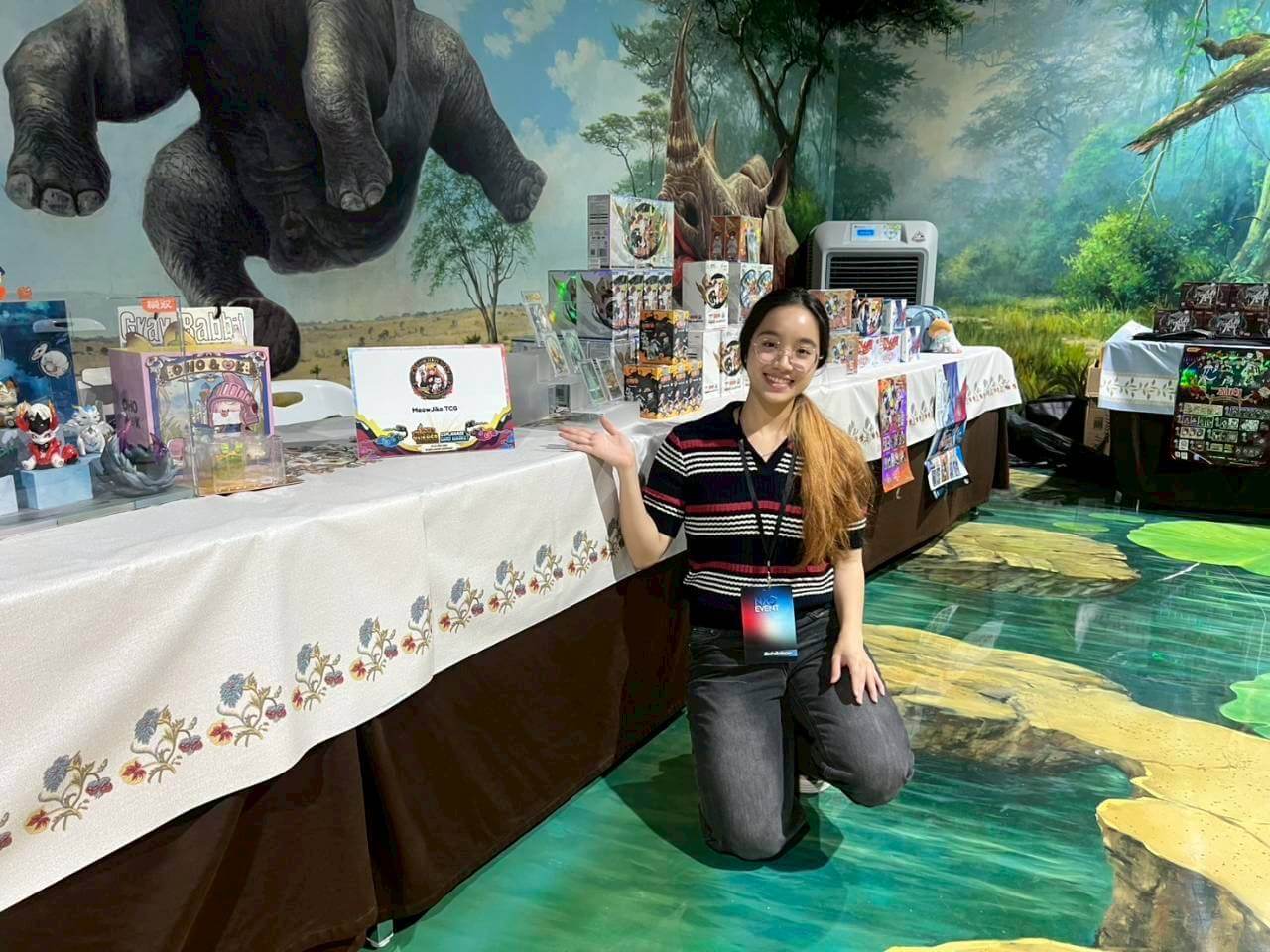 A woman kneeling in front of a display table at what appears to be a themed event or convention, with a large illustrated gorilla mural in the background and merchandise arranged on tables.