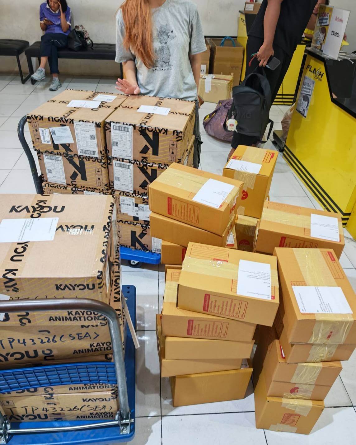 Multiple cardboard shipping boxes, some branded KAYOU and labeled with shipping labels, are stacked on a blue cart and the floor in what appears to be a warehouse or sorting area. A person in a light gray graphic T-shirt stands behind the boxes, and another person is partially visible near a yellow counter on the right.